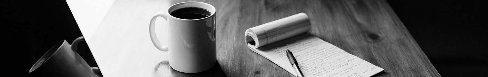 A photo of a coffee mug, pad of paper, and pen sitting on a desk.